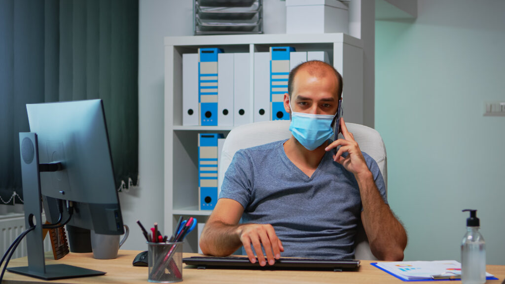 Man wearing a face mask talking on the phone in office while working on ACA health insurance tasks.
