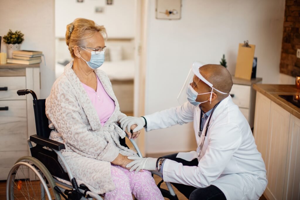Doctor wearing face shield and mask talking to senior woman in wheelchair during home visit under Affordable Care Act Indiana coverage.